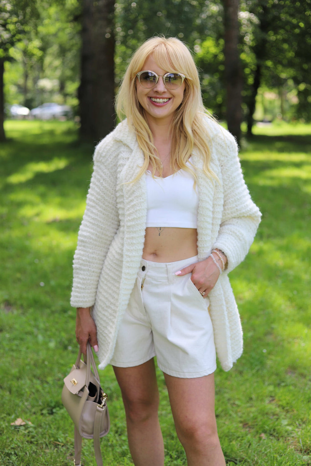Uśmiechnięta kobieta w parku w ręcznie robionej marynarce Coco oversize z alpaki Sirka w kolorze śmietankowym