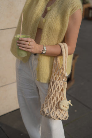 Ręka trzymająca kawę i torebkę dzierganą na szydełku z brelokiem krewetka w odcieniu masełkowym Sirka handmade