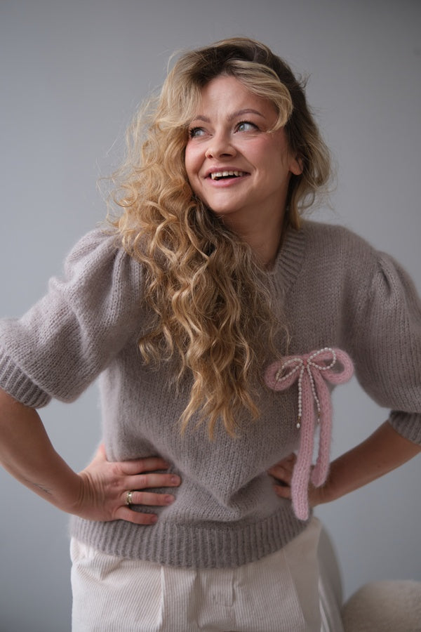 Uśmiechnięta kobieta w swetrze z broszką w kształcie kokardki z perłami.
