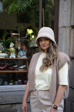 Stojąca kobieta w kapeluszui kamizelce w odcieniu ciepły beż Sirka handmade na tle kawiarni
